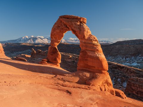 Delicate Arch glowing orange at sunset with snow-capped mountains visible through the opening.