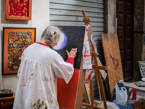 An artist in a paint-stained smock working at an easel in a small studio surrounded by finished canvases.