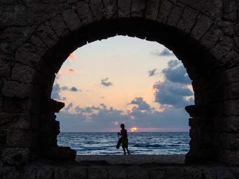 A figure silhouetted on a beach, framed by the stone arch of an ancient aqueduct ruin at sunset.