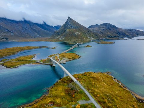 Aerial view of a curving bridge crossing turquoise water between small autumn-colored islands, with a pointed mountain rising into low clouds.