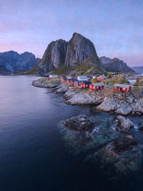 Red fishermen's cabins on stilts beneath a towering granite peak, with pink and lavender twilight clouds above a dark fjord.
