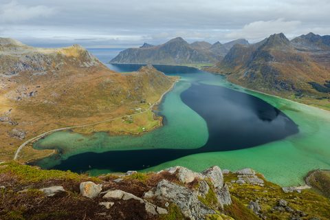 View from a mountainside down to a fjord where turquoise shallows meet a deep navy channel, ringed by autumn-colored mountains.