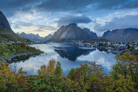 A jagged, cloud-capped mountain reflected in a still fjord beside a fishing village, framed by autumn foliage and red rowan berries.