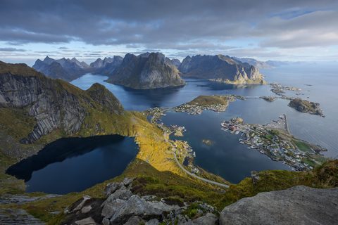 Elevated view from a mountain ridge over fjords, dark lakes, and island villages connected by bridges, with sharp peaks under a dramatic cloudy sky.
