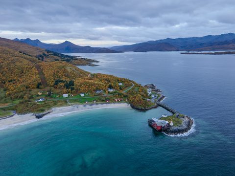 Aerial view of a sandy beach and turquoise bay below autumn birch hillsides, with a red pier building on a rocky breakwater and mountains beyond.