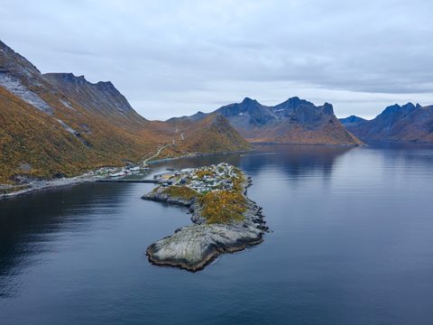 Aerial view of a small village on a rocky peninsula in a calm fjord, with autumn-colored slopes and blue mountains behind.