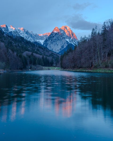 Snow-capped mountain peaks lit pink by alpenglow above a calm lake at dawn.