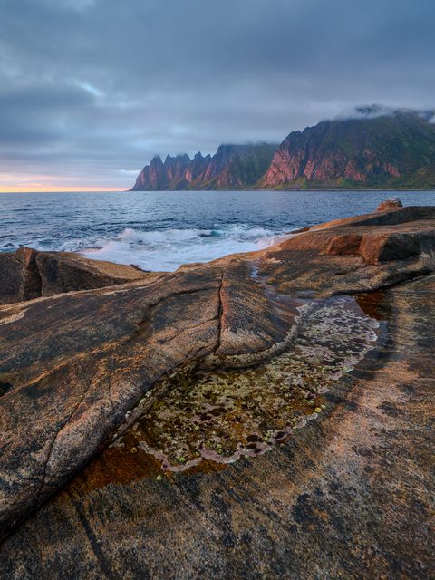 Weathered coastal rock with tide pools in the foreground and jagged mountain peaks along the shoreline at dusk.
