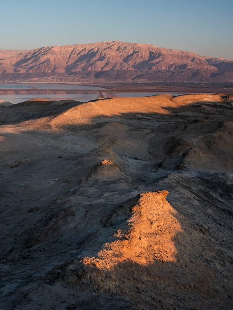Sunlit desert badlands above the Dead Sea, with distant mountains and the sea visible in the haze.